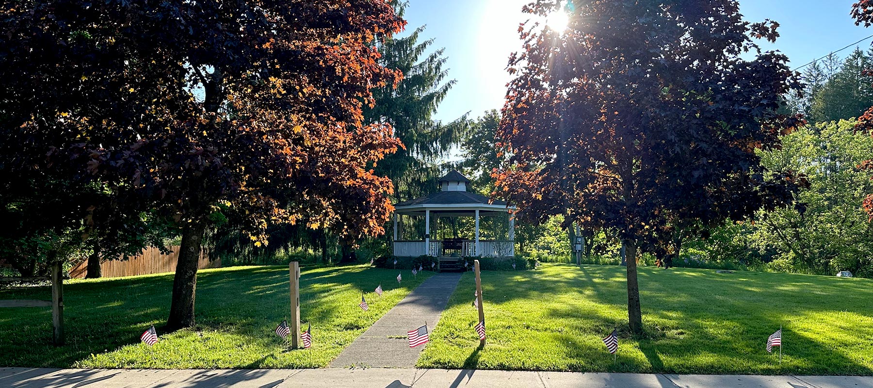 Photo of a bandstand on green grass with trees and the sun setting behind.